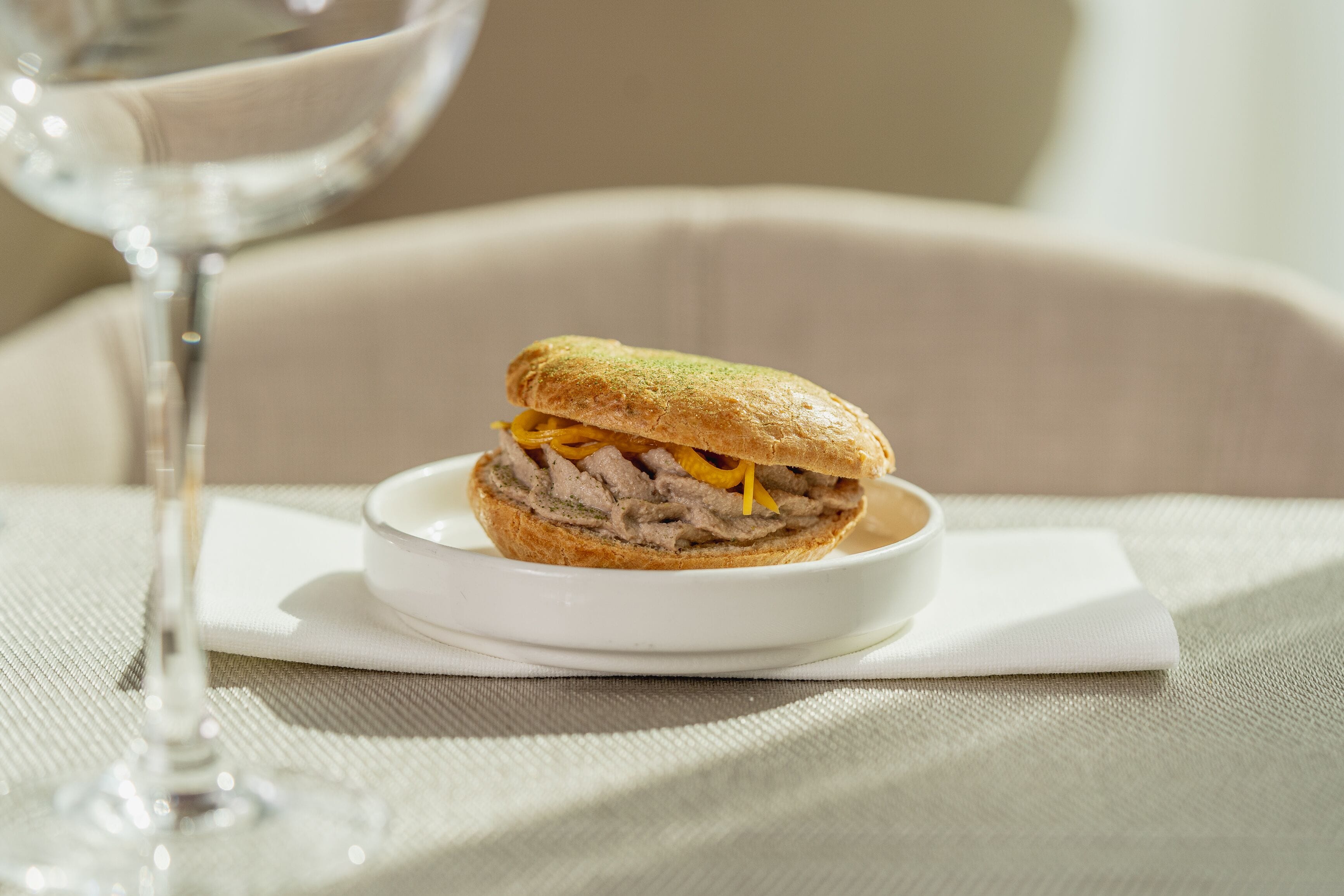 A sandwich on a white plate with a napkin, a glass of water, and a chair in the background.