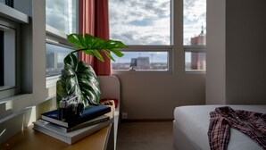 Una habitación de hotel con una cama, un escritorio con libros y una cámara, una planta en maceta y una ventana con vista a la ciudad.
