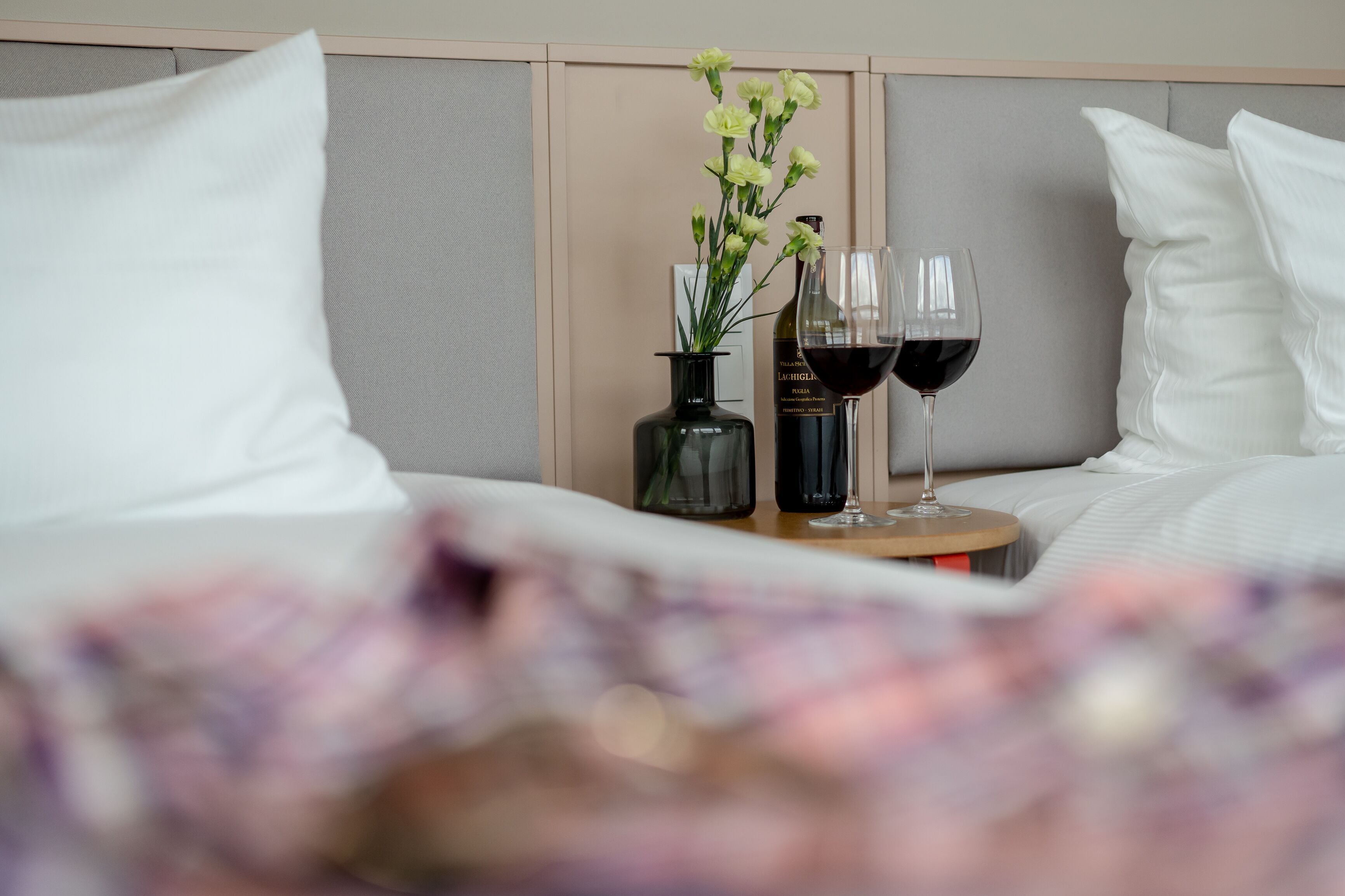 Cama bem arrumada com almofadas brancas e cabeceira, uma pequena mesa com um vaso de flores e dois copos de vinho tinto.