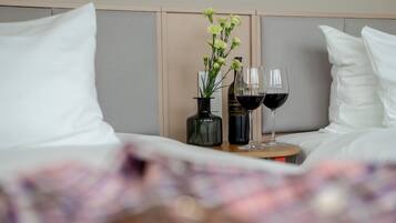 A neatly made bed with white pillows and a headboard, a small table with a vase of flowers and two wine glasses filled with red wine.