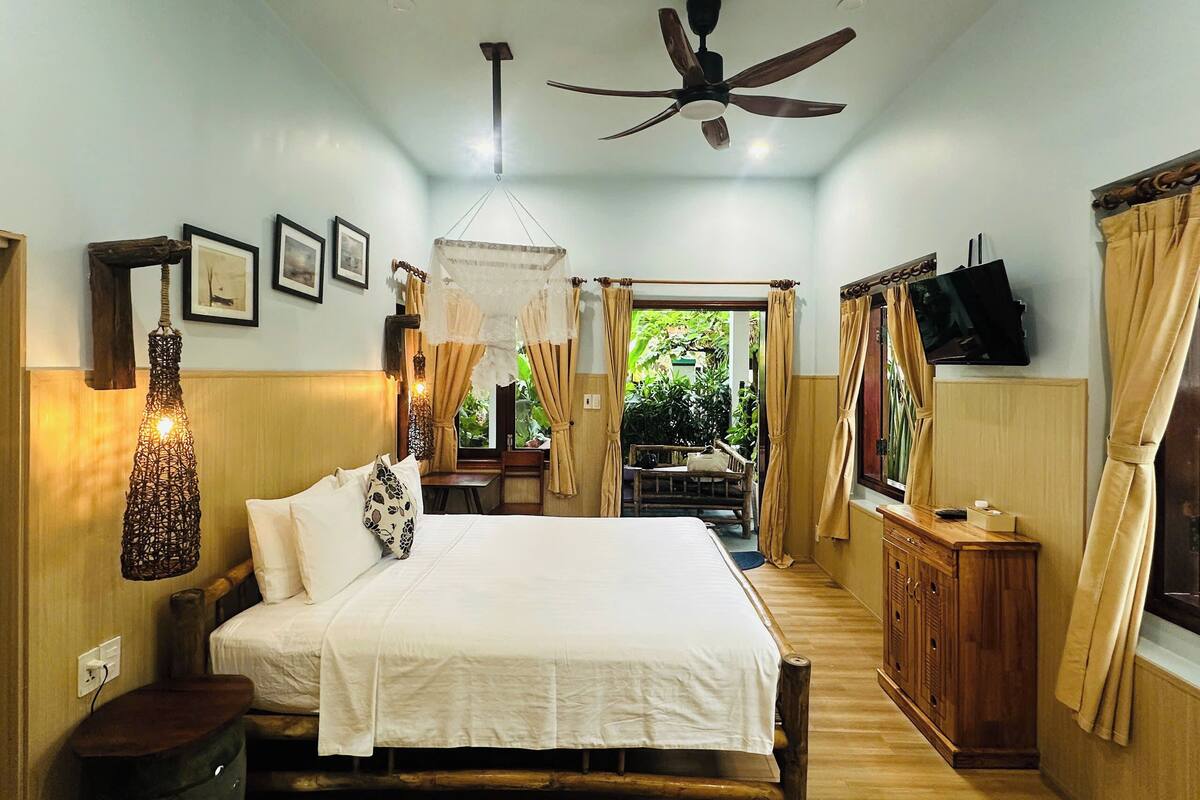 A bedroom with a bed, bedside table, hanging light fixture, ceiling fan, and a view of the outdoors.