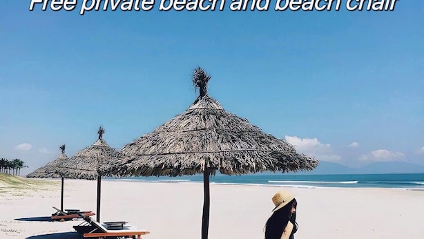 Una playa con sombrillas de paja y sillas de descanso, una mujer con vestido a rayas y un cielo azul despejado.