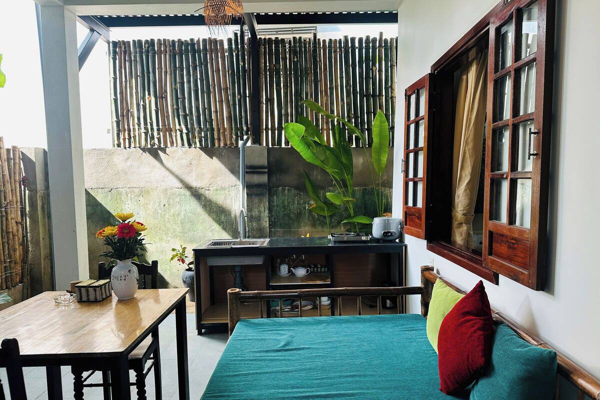 A room with a bed, a table, and a bench. There is a window with curtains and a bamboo fence outside.