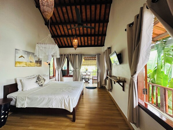 A bedroom with a wooden floor, a bed with white bedding, a ceiling fan, a wall-mounted TV, and a window with curtains.