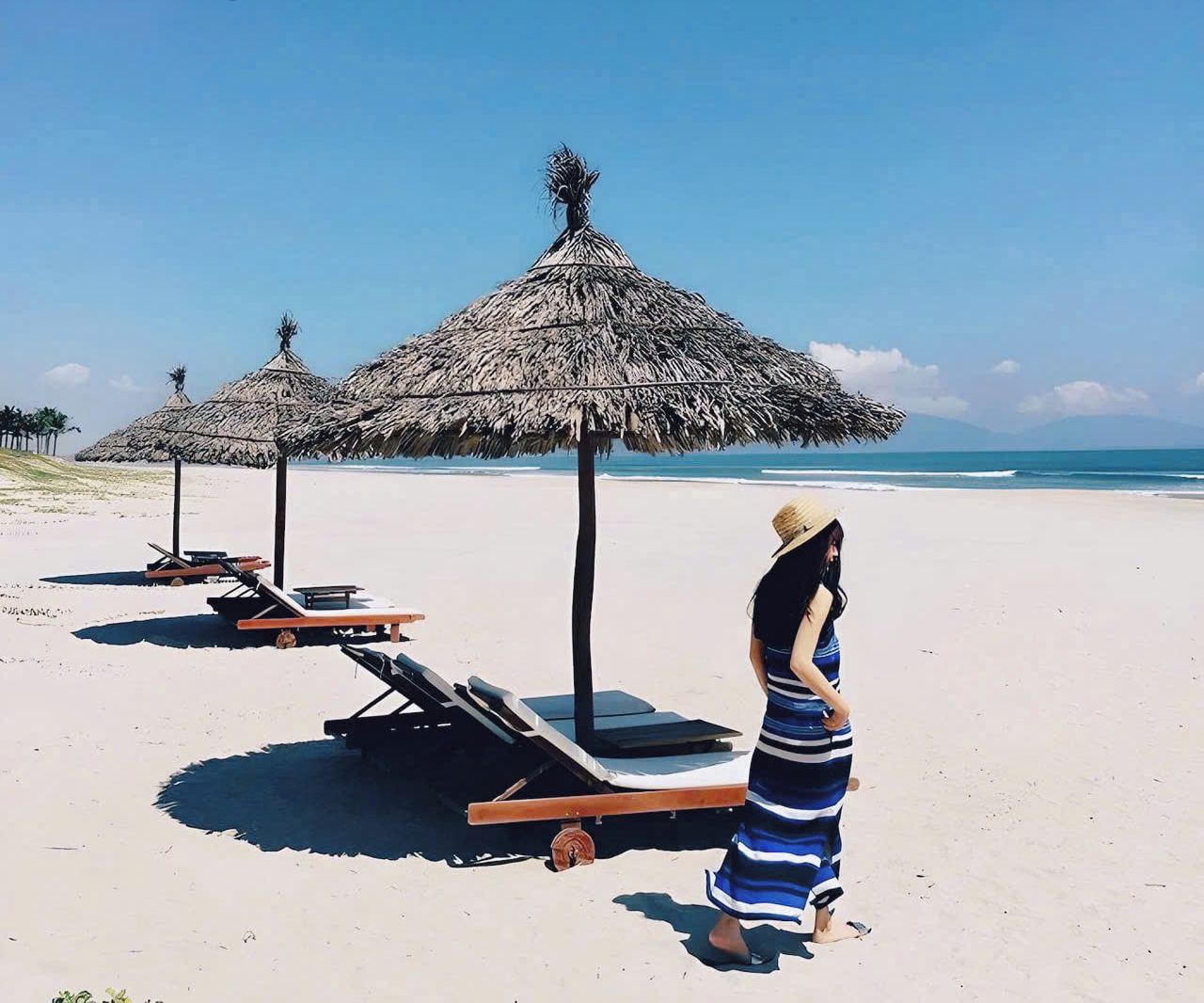 A beach with thatched umbrellas and lounge chairs, a woman in a striped dress, and a clear blue sky.