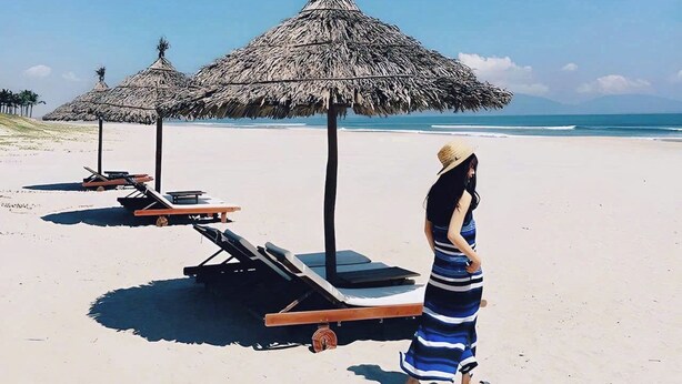 Une plage avec des parasols en chaume et des chaises longues, une femme portant une robe à rayures et un ciel bleu dégagé.