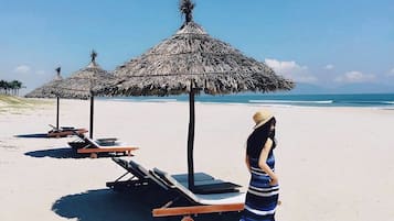A beach with thatched umbrellas and lounge chairs, a woman in a striped dress, and a clear blue sky.