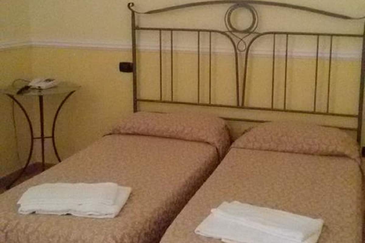 A room with two beds, each with a beige bedspread and pillows, a small round table with a lamp, and a wall-mounted light fixture.