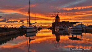 Un puerto deportivo con barcos amarrados, un molino y un edificio reflejado en aguas tranquilas durante el atardecer.