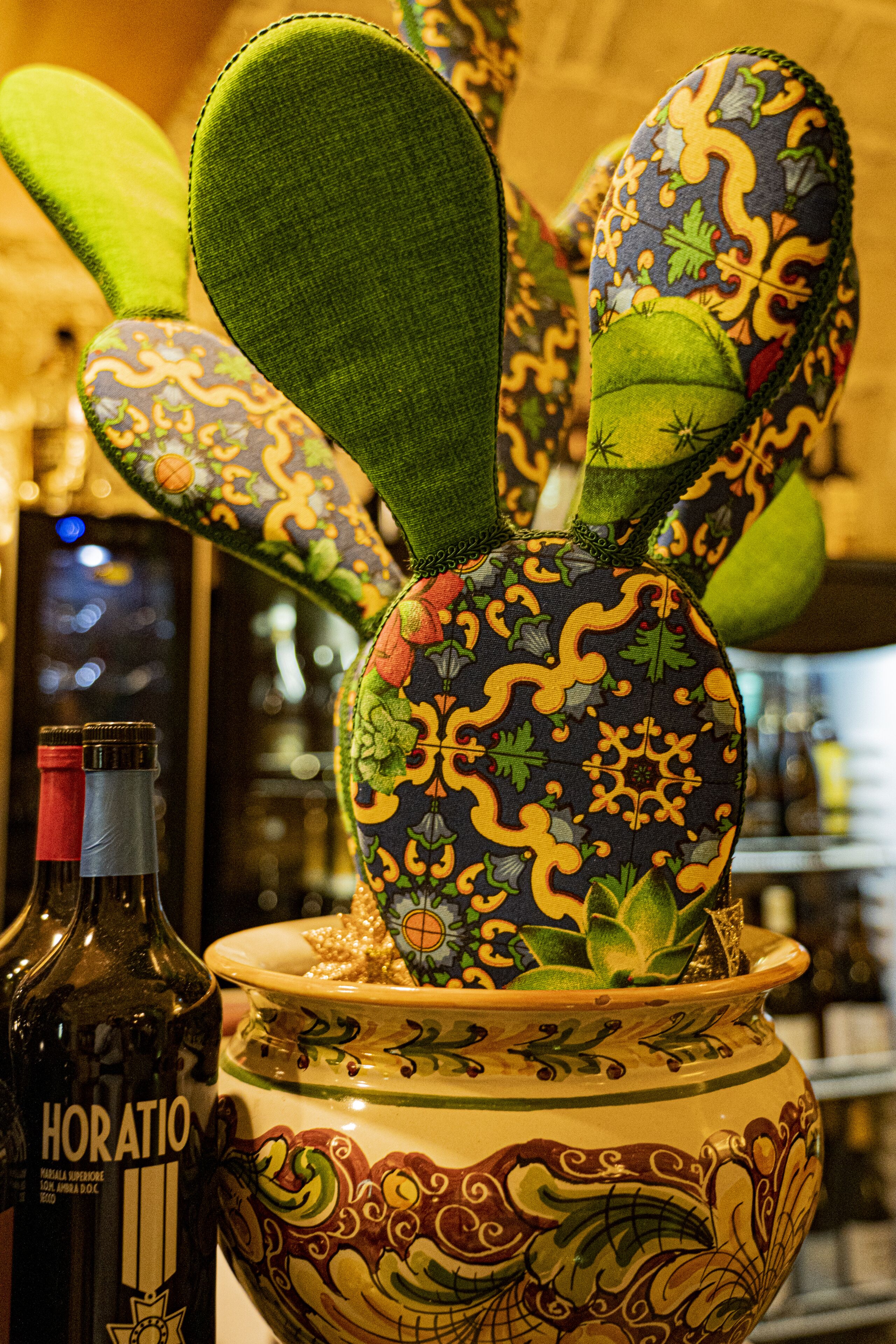 A cactus sculpture with a patterned pot, a bottle of Horatio wine, and a blurred background of shelves.
