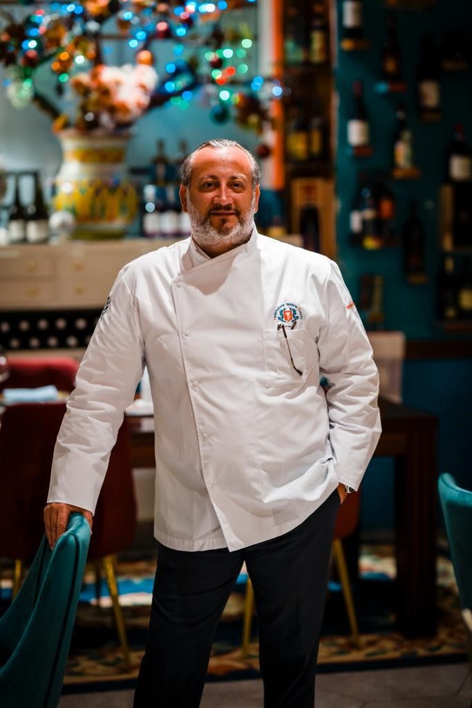A person in a white chef's coat standing in a room with a bar and chairs.