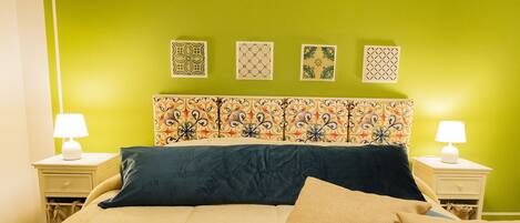 Een bed met een beige en blauw hoofdeinde, twee bedlampjes en vier decoratieve wandhangers.