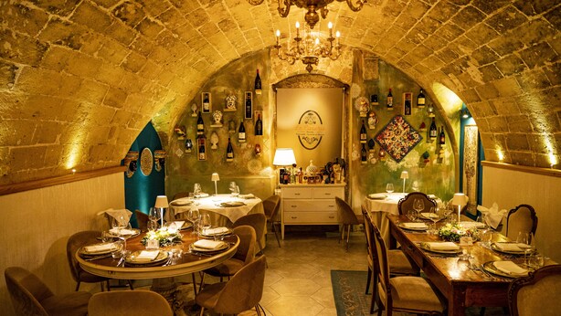 Un comedor con techo abovedado, una araña y una pared decorada con botellas de vino y un reloj.