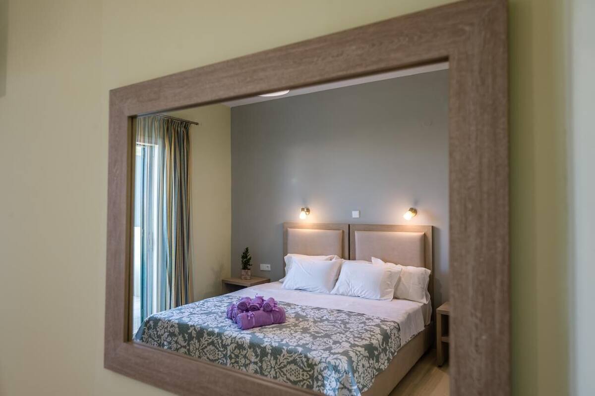 Una habitación de hotel con un espejo grande, una cama con sábanas blancas, una mesita de noche pequeña con una lámpara y una ventana con cortinas.