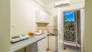 A compact kitchen with a built-in stove, a small sink, and a cabinet. A balcony with a metal railing is visible through an open door. - Blue Lagoon (Zakynthos)