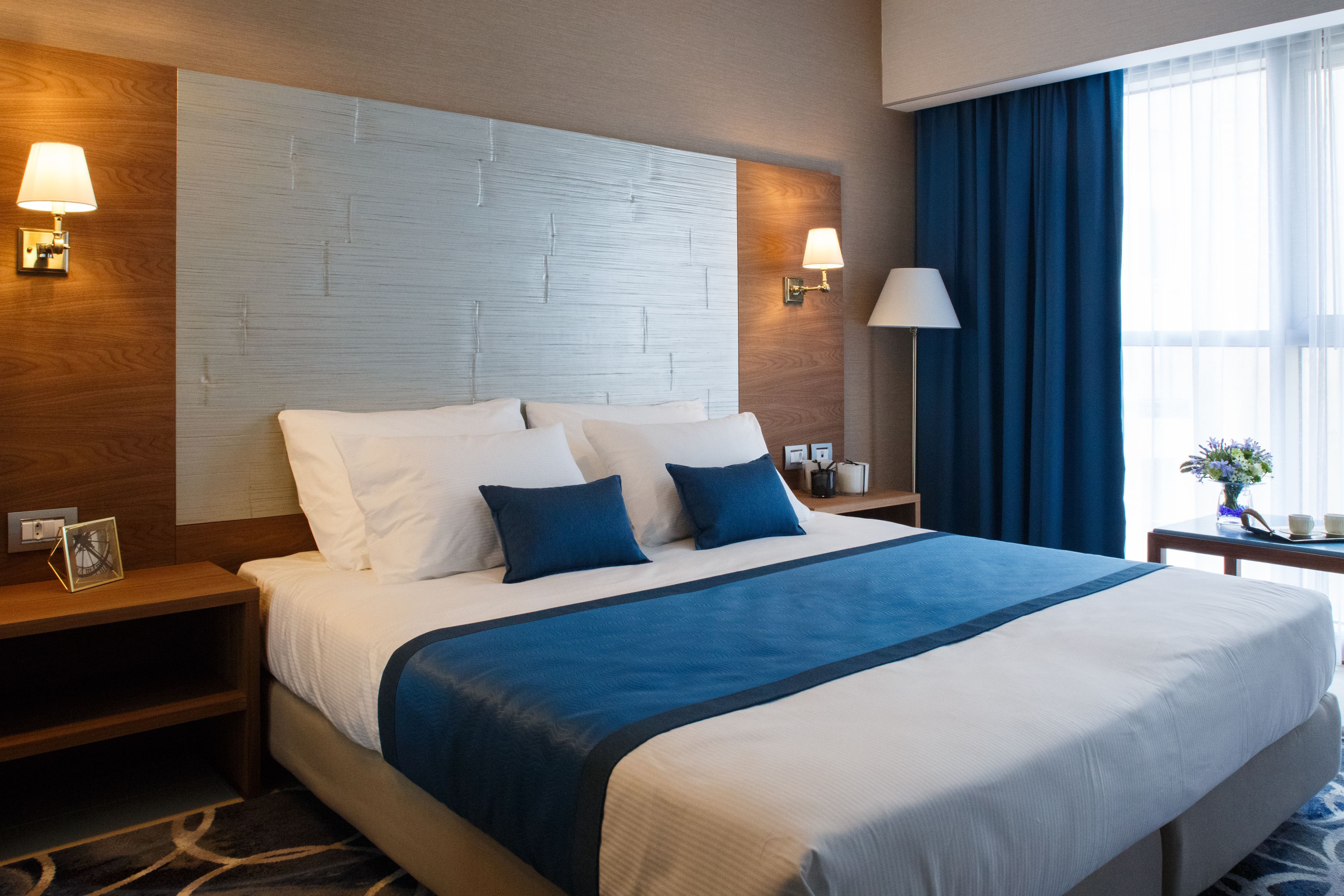 Une chambre d’hôtel avec un grand lit, deux tables de chevet, une petite table, une lampe et une fenêtre avec des rideaux.