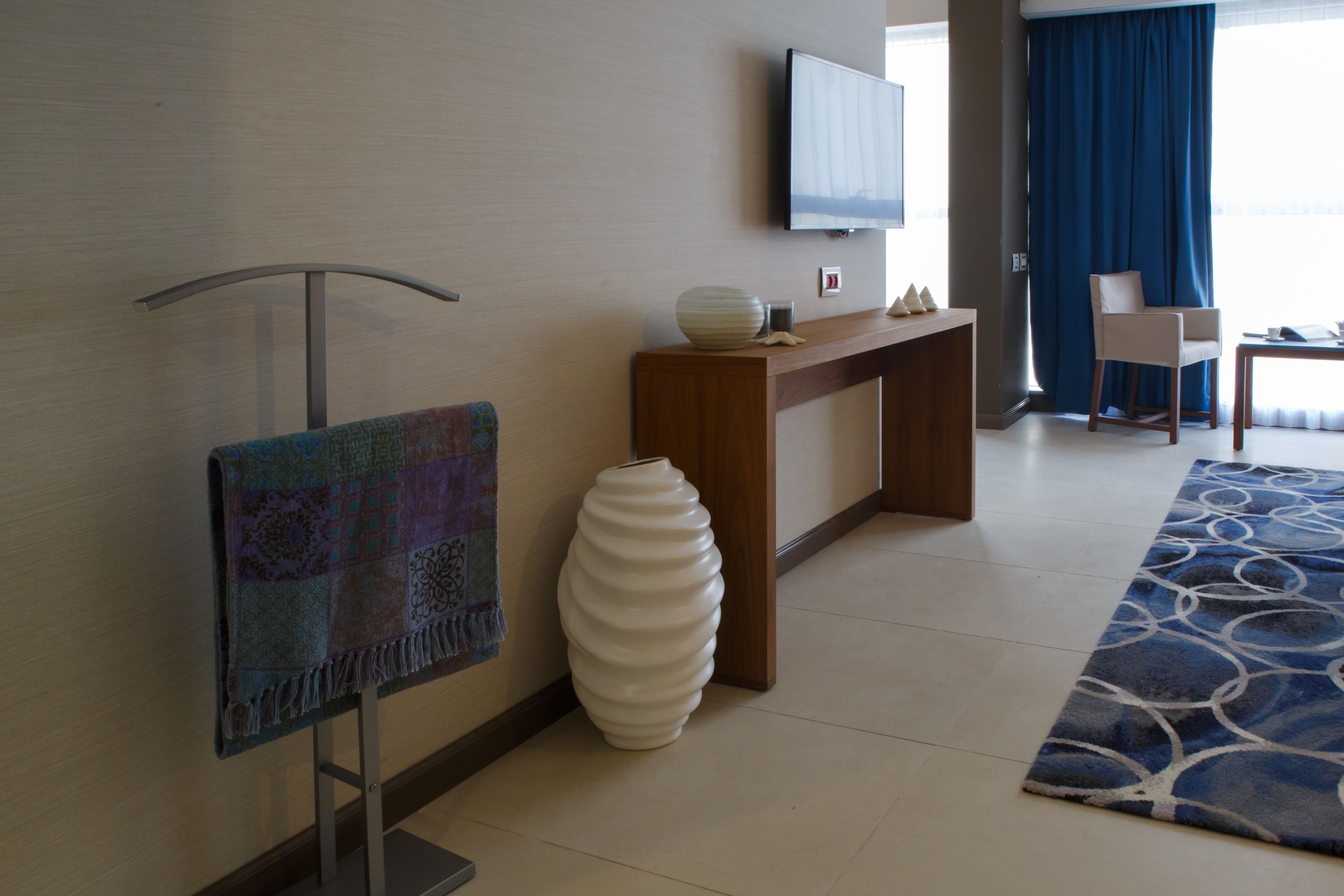 A modern hotel room with a flat-screen TV mounted on the wall, a wooden console table, a white vase, a colorful tapestry, and a blue patterned rug.