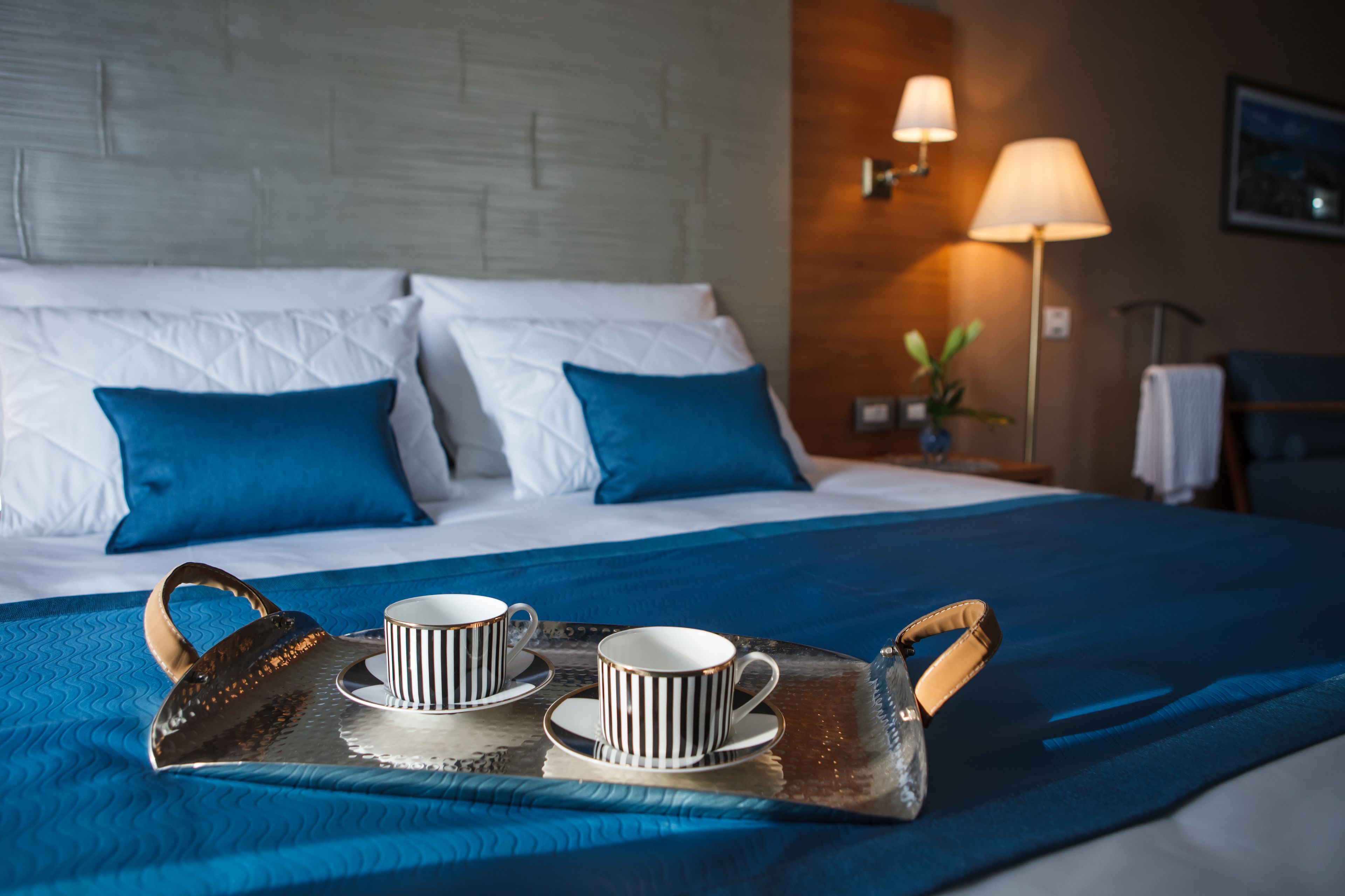 A neatly made bed with blue and white bedding, a tray with two cups and a teapot, and a lamp on the side.