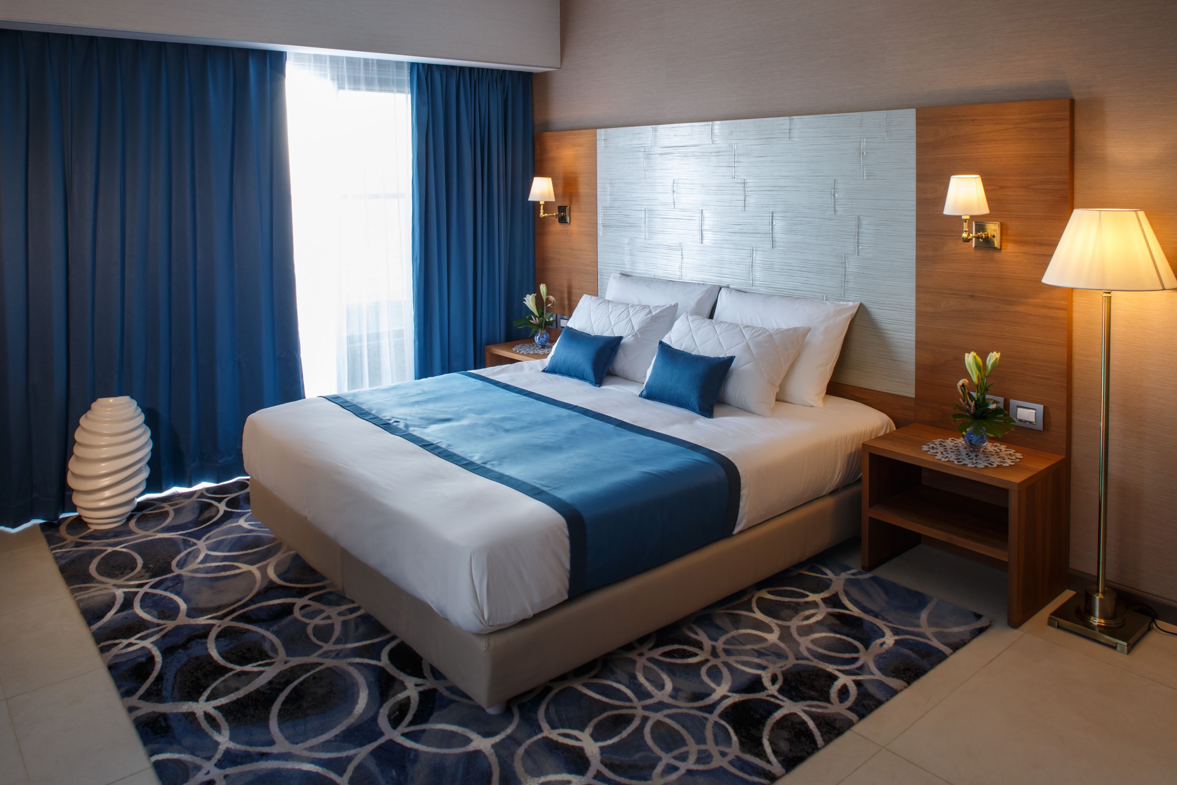 A modern hotel room with a large bed, blue and white bedding, two bedside lamps, a nightstand with a vase, and a patterned rug.