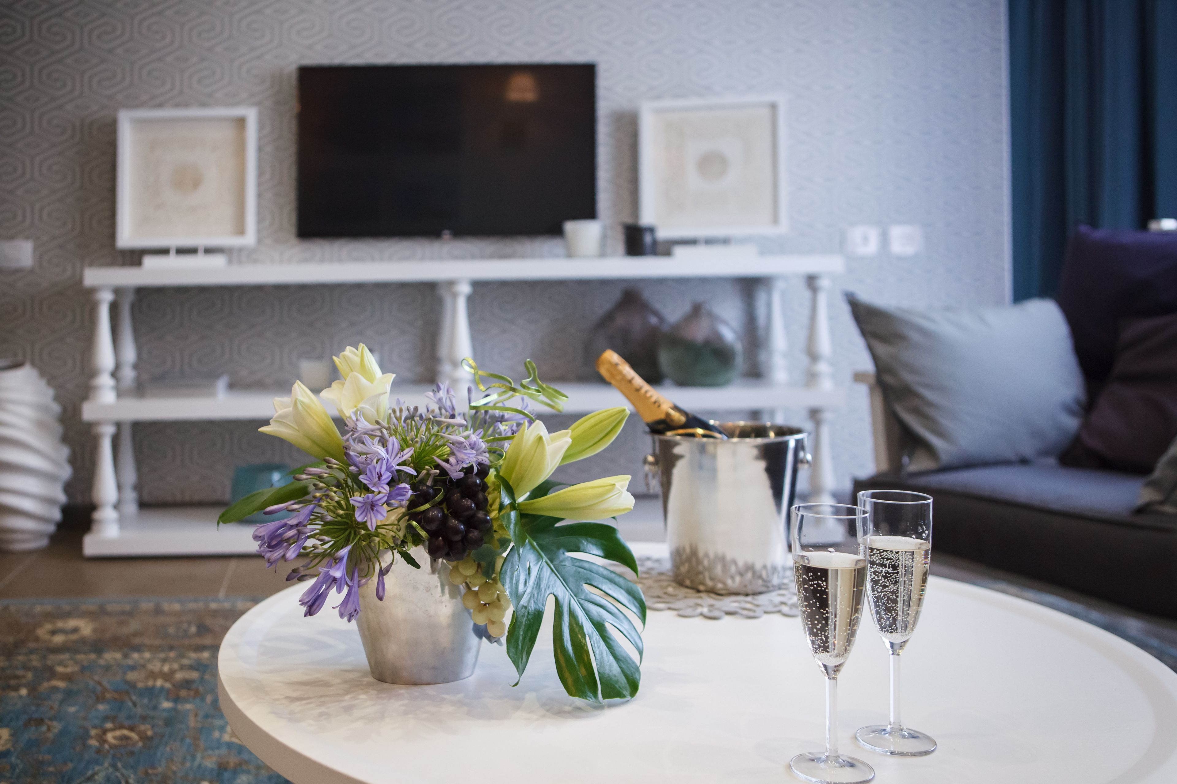 A living room with a round coffee table, a floral centerpiece, two champagne glasses, and a TV on the wall.