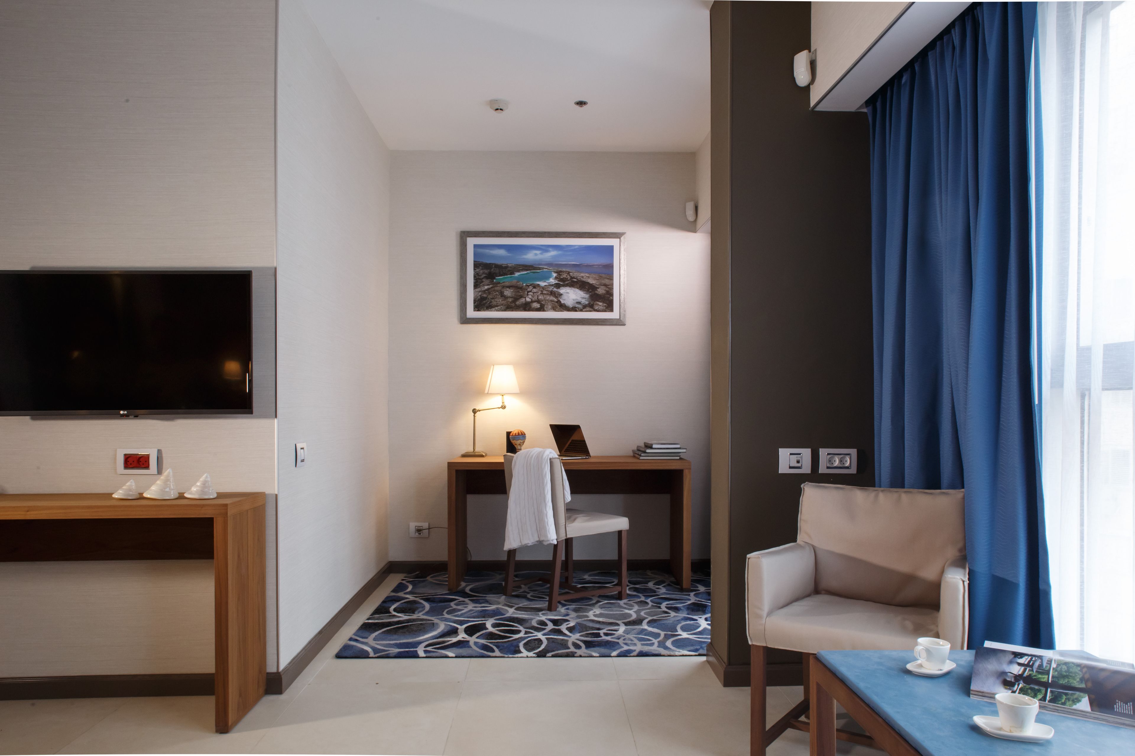 A hotel room with a TV, a desk with a lamp, a chair, a sofa, and a window with curtains.