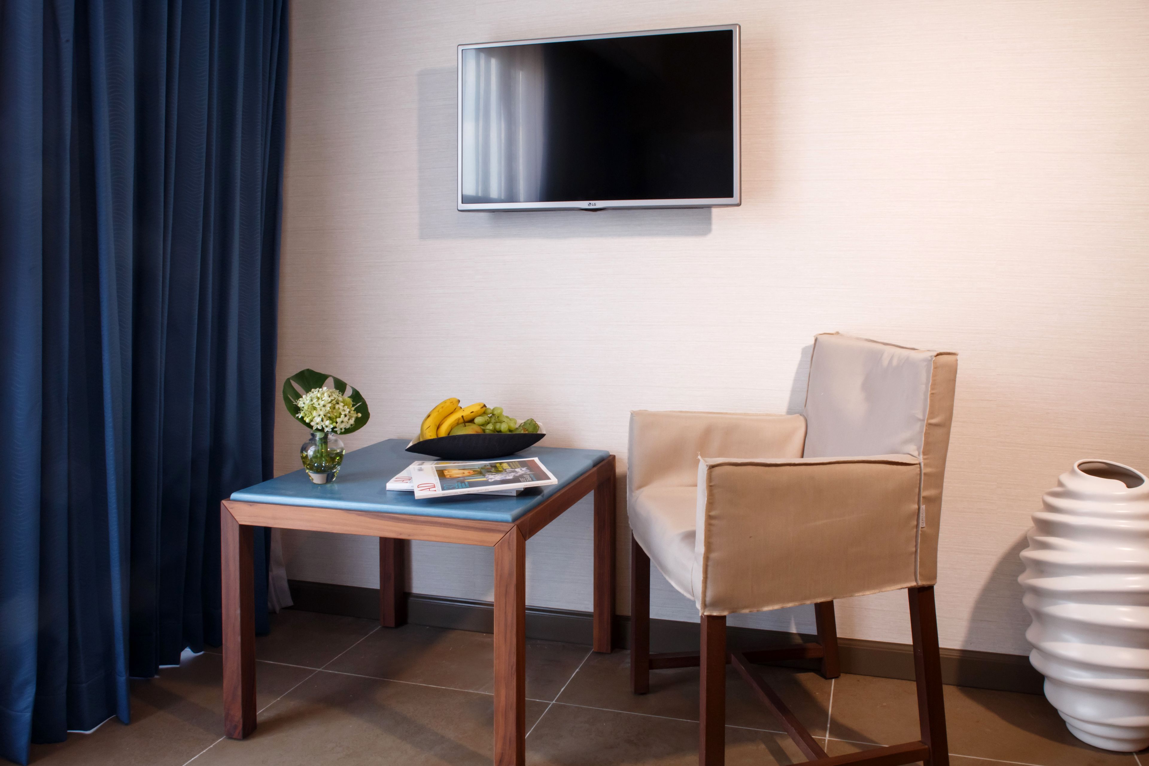A hotel room with a flat-screen TV mounted on the wall, a wooden table with a bowl of fruit and a vase, a beige armchair, and a white vase on the floor.