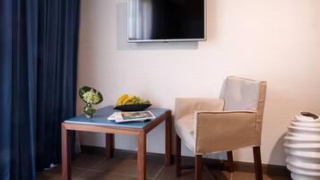 A hotel room with a flat-screen TV mounted on the wall, a wooden table with a bowl of fruit and a vase, a beige armchair, and a white vase on the floor.
