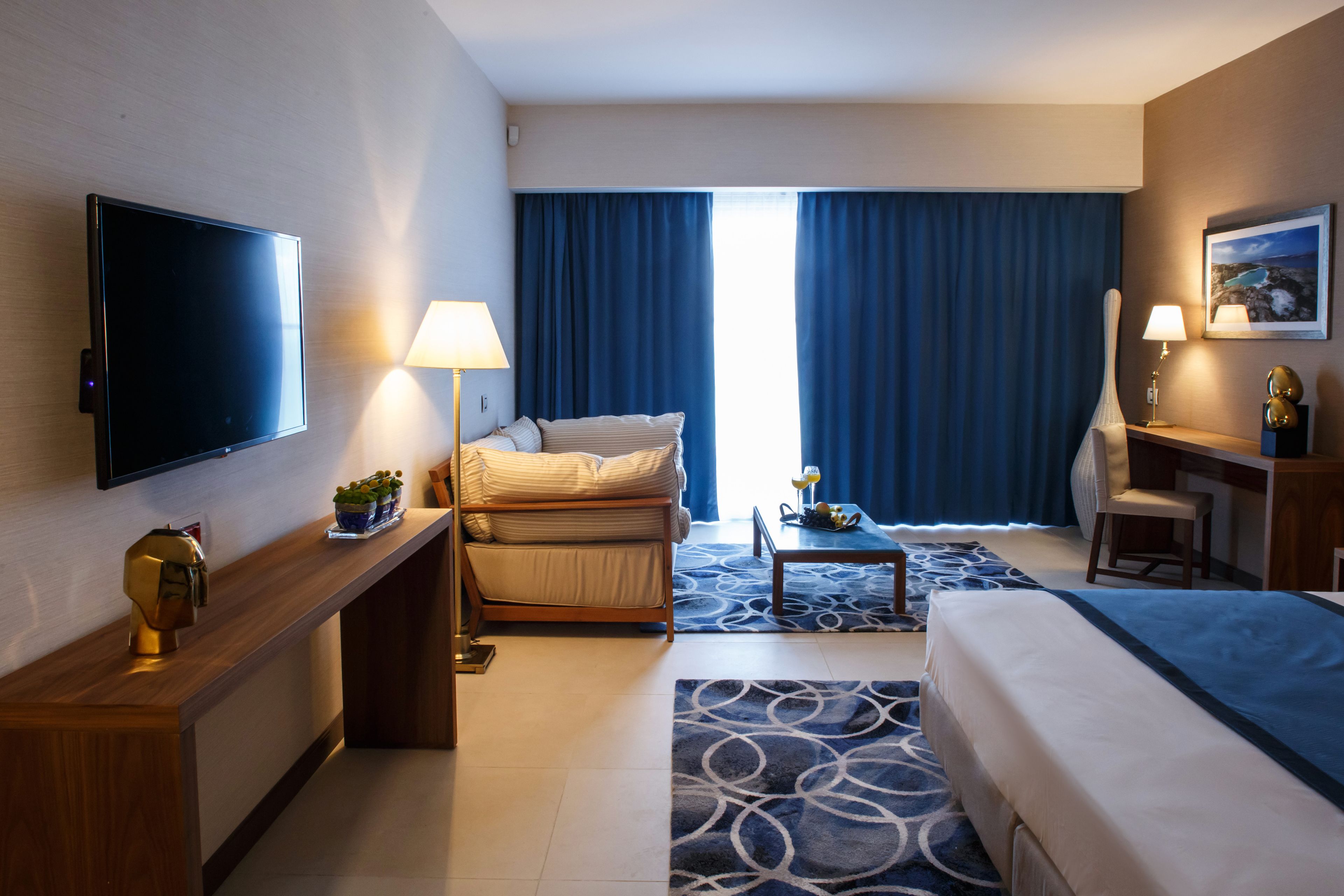 A hotel room with a flat-screen TV, a sofa, a desk with a chair, a bed, and a large window with curtains.