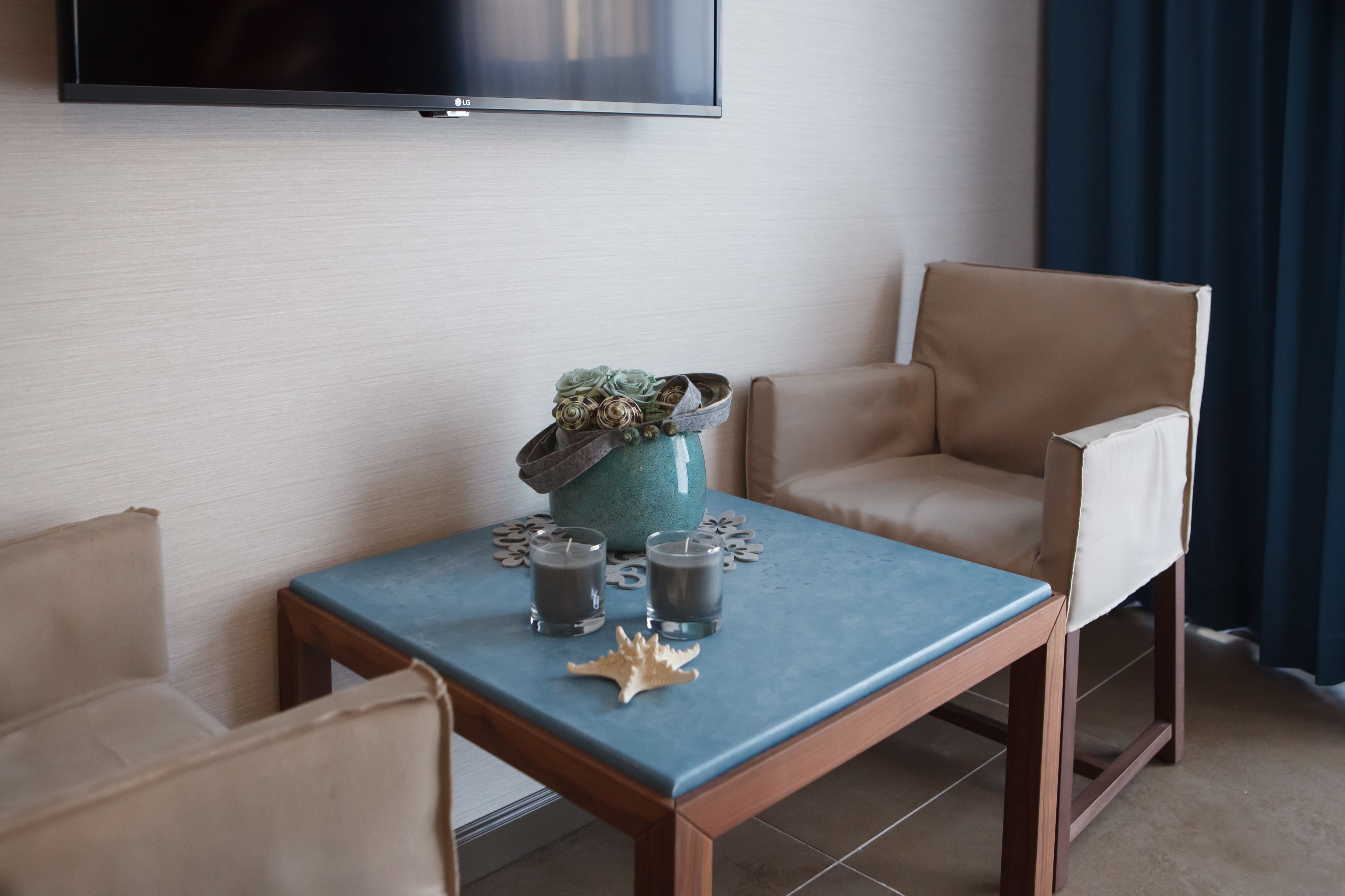 A hotel room with a flat-screen TV mounted on the wall, a small table with two candles and decorative items, and two beige armchairs.