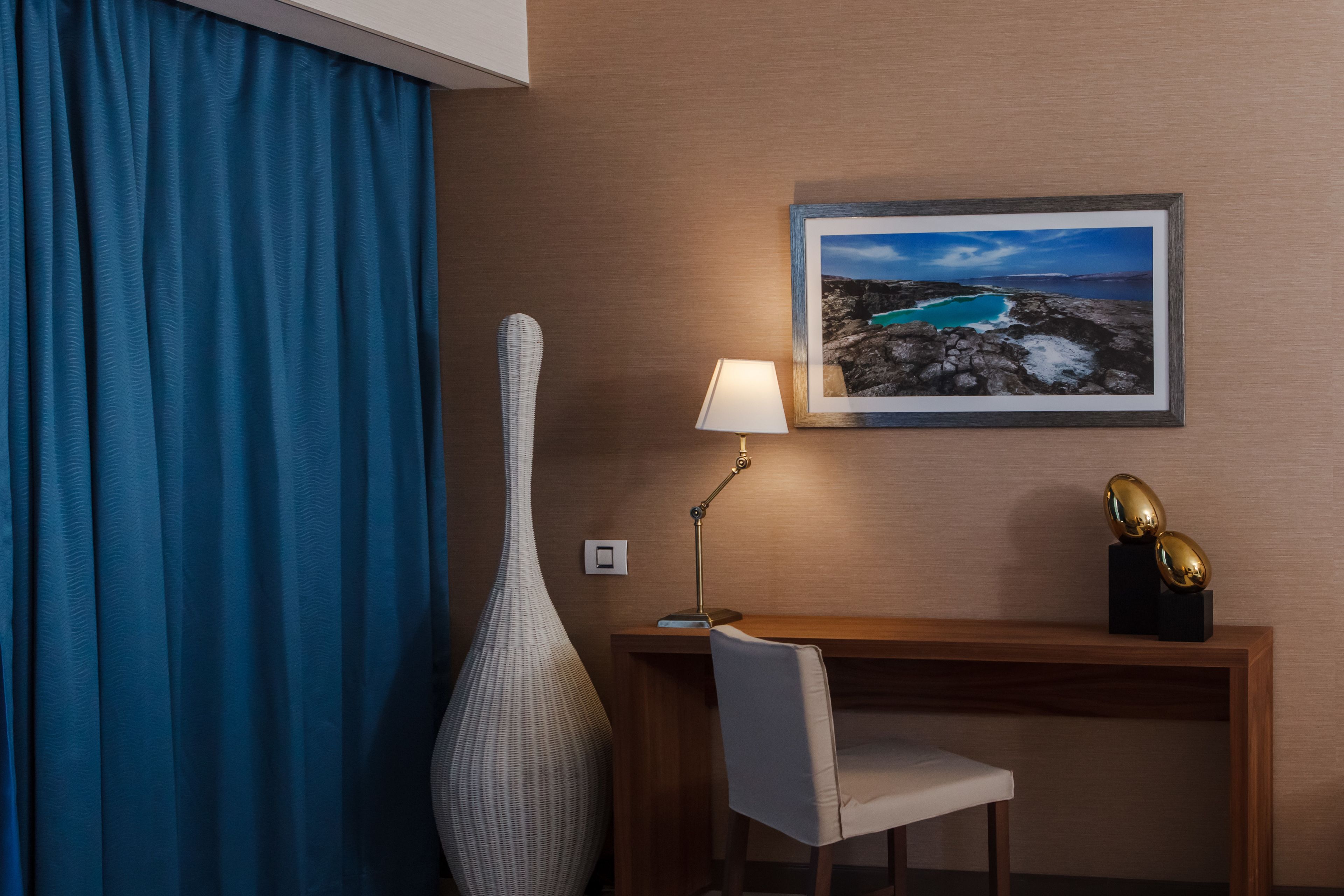 A hotel room with a desk, chair, lamp, large vase, and a framed picture on the wall.