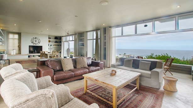 A spacious living room with a large window overlooking the sea, furnished with leather sofas, a coffee table, and a rug. - 10 Elf Beach House (Glentana)