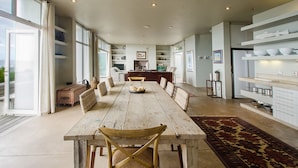 A spacious dining area with a long wooden table, surrounded by wicker chairs, and a kitchen area with white cabinets and shelves. - 10 Elf Beach House (Glentana)