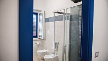 A modern bathroom with a glass shower enclosure, two toilets, and a window with blinds.