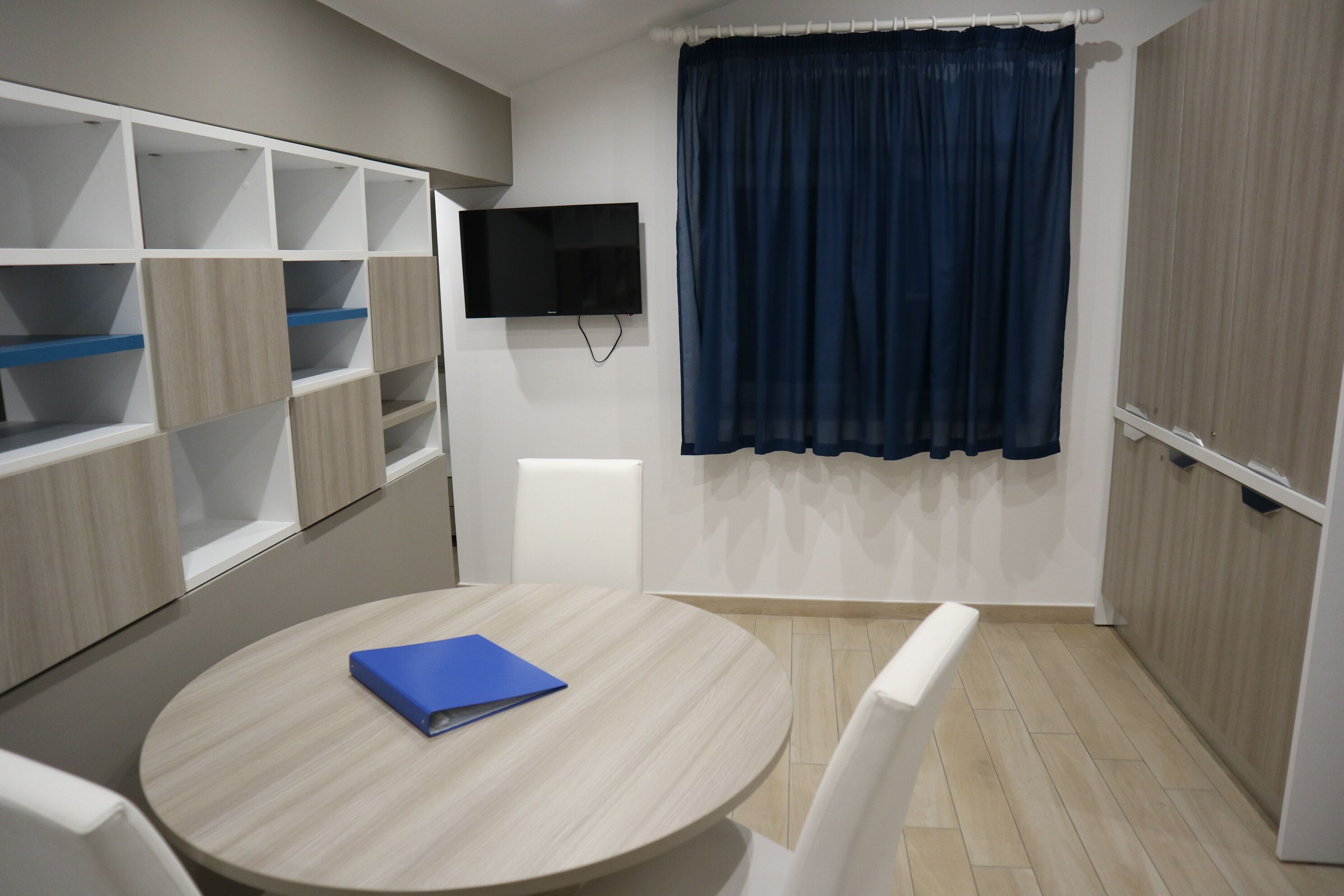 Une pièce avec une table ronde, un classeur bleu, un téléviseur accroché au mur et un rideau bleu.