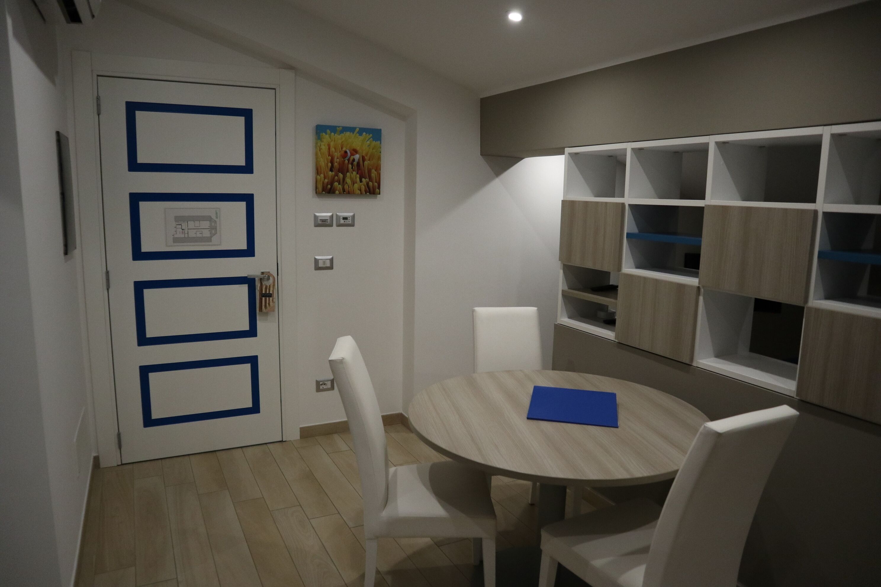 Une pièce moderne avec une table ronde en bois, des chaises blanches, une porte blanche avec des encadrements bleus et des étagères intégrées aux finitions en bois clair.