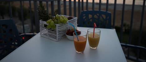 Una mesa con dos bebidas y una cesta de frutas en un balcón con vistas al mar al atardecer.