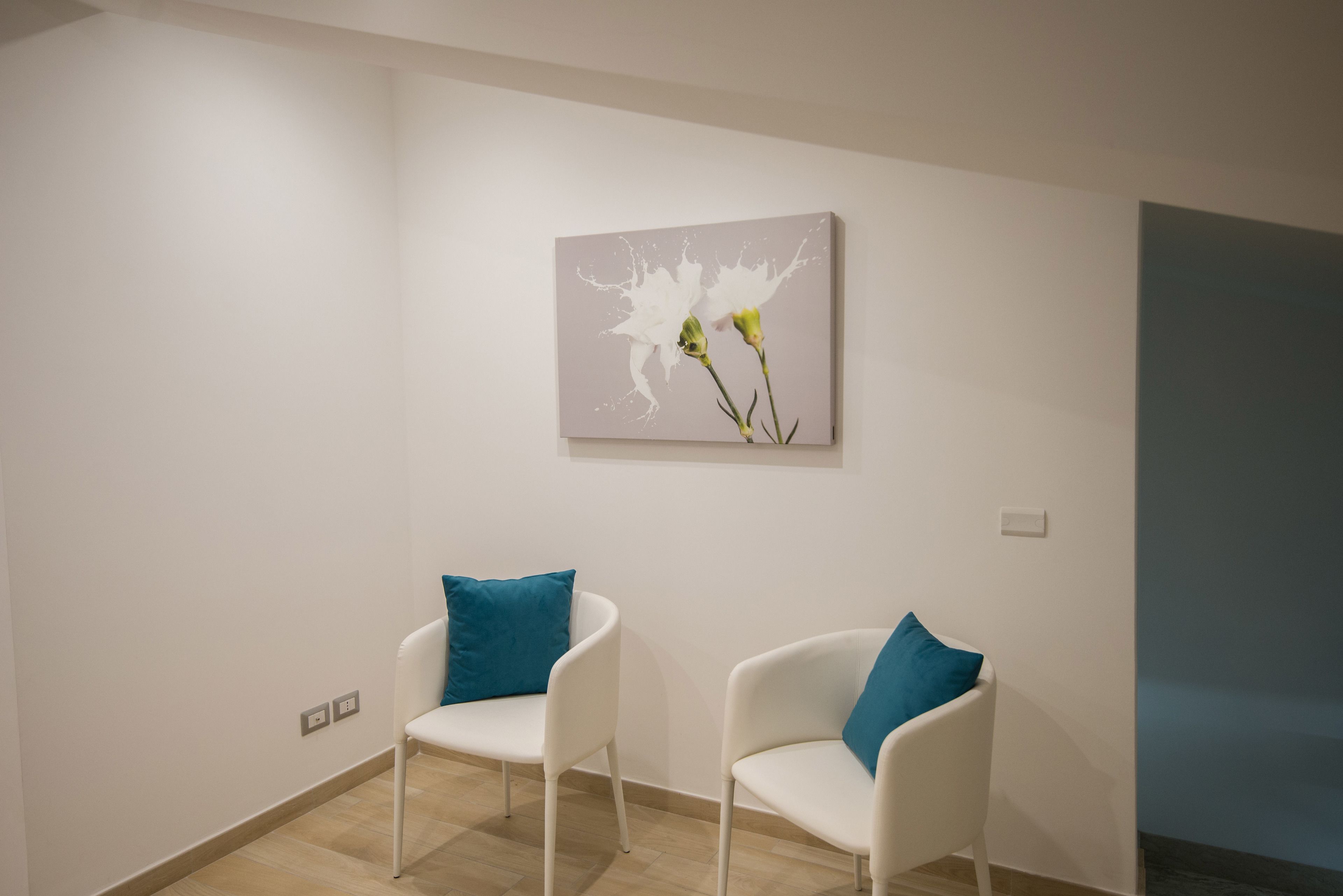 Een moderne kamer met twee witte stoelen, elk met een blauwgroen kussen, en een kunstwerk aan de muur met bloemen.