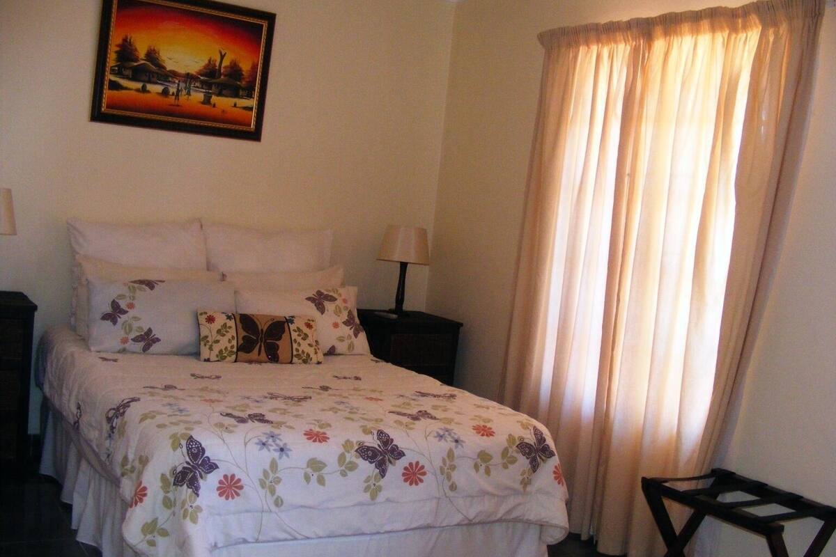 A bedroom with a bed, pillows, a nightstand, a lamp, a chair, and a painting on the wall.