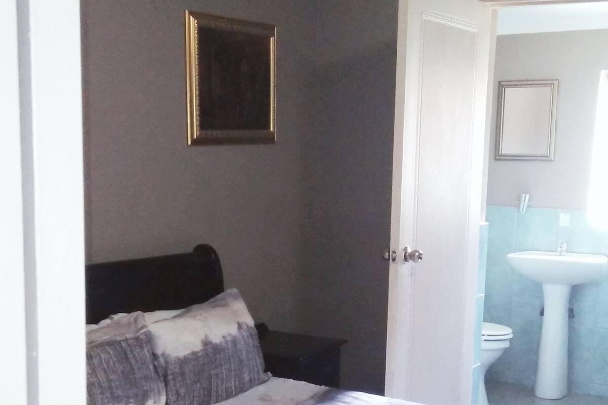 A bedroom with a bed, a nightstand, a framed picture on the wall, and a bathroom with a sink and toilet visible through an open door.