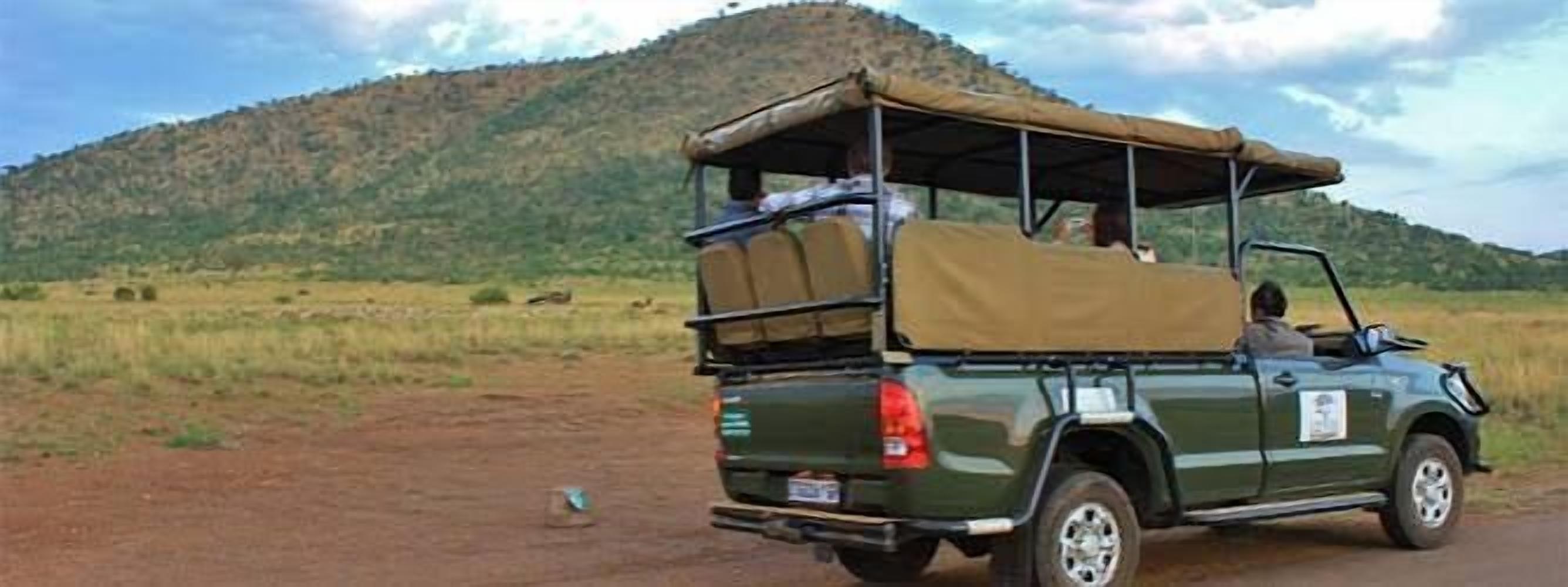 A green off-road vehicle with a roof and luggage on the back, parked on a dirt road with a hilly landscape in the background.