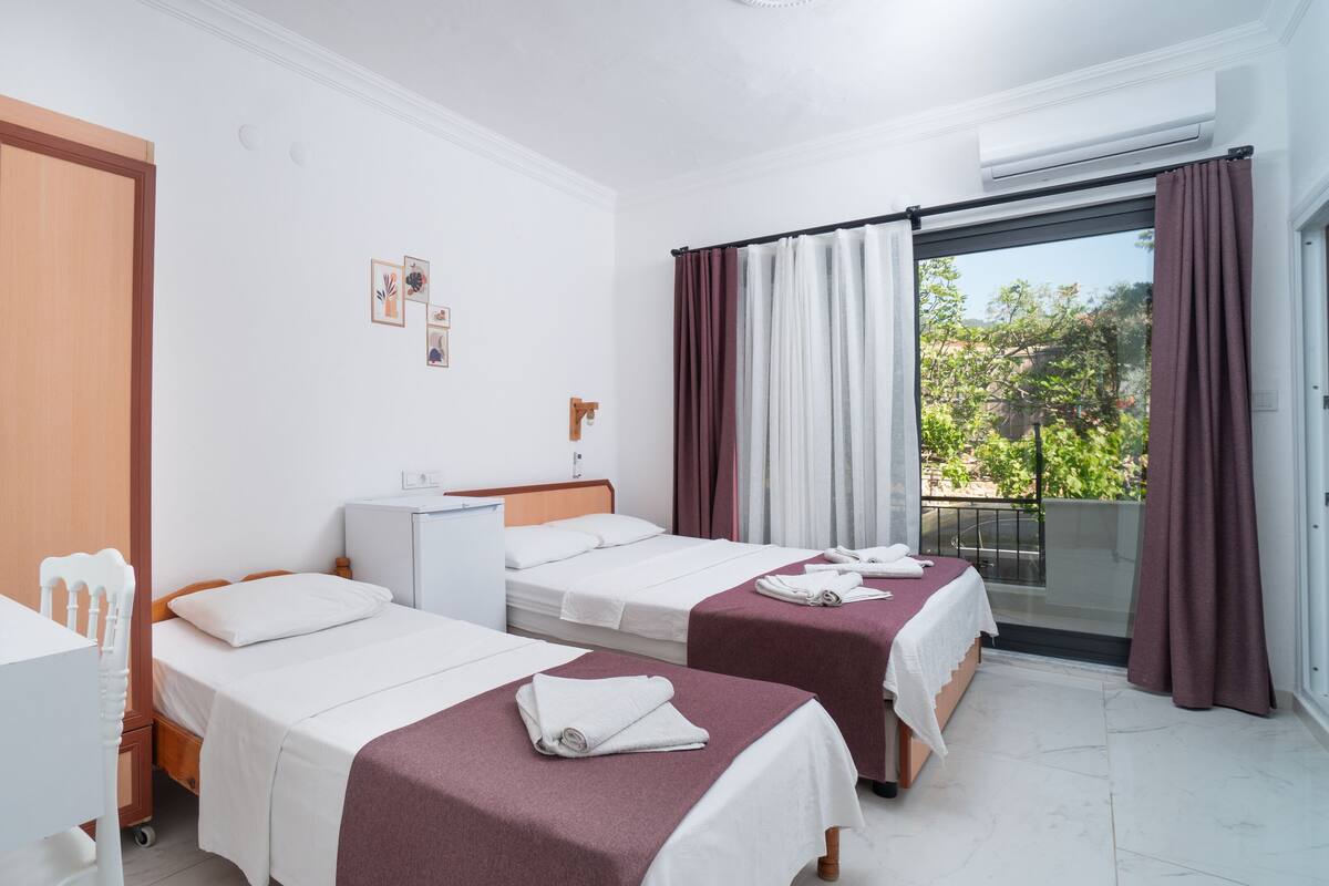 Habitación de hotel con dos camas individuales, un escritorio pequeño, un refrigerador y puerta corrediza de vidrio a un balcón.