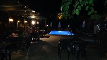 An outdoor dining area with tables and chairs, a pool, and umbrellas providing shade.