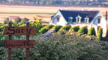 A wooden sign with "Sacy Champagne 1er Cru" written on it, in front of a house with a blue roof and white walls, surrounded by greenery.