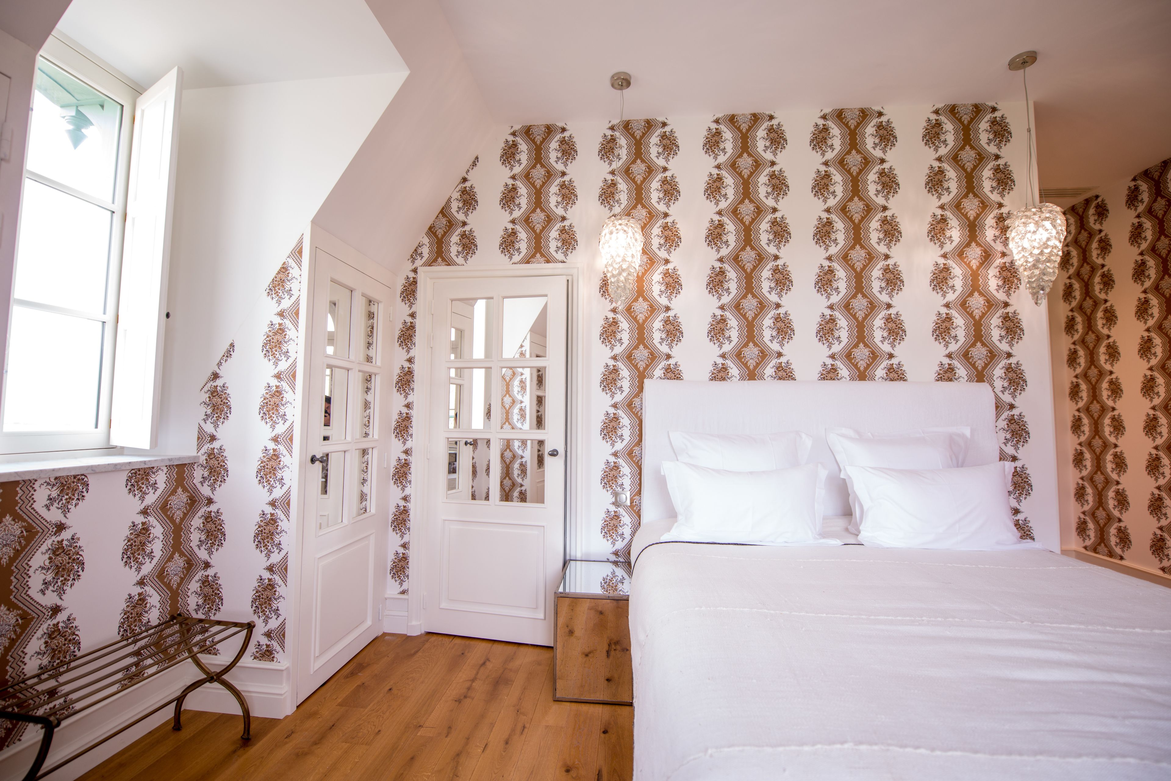 A bedroom with a bed, a bench, a door, and patterned wallpaper.