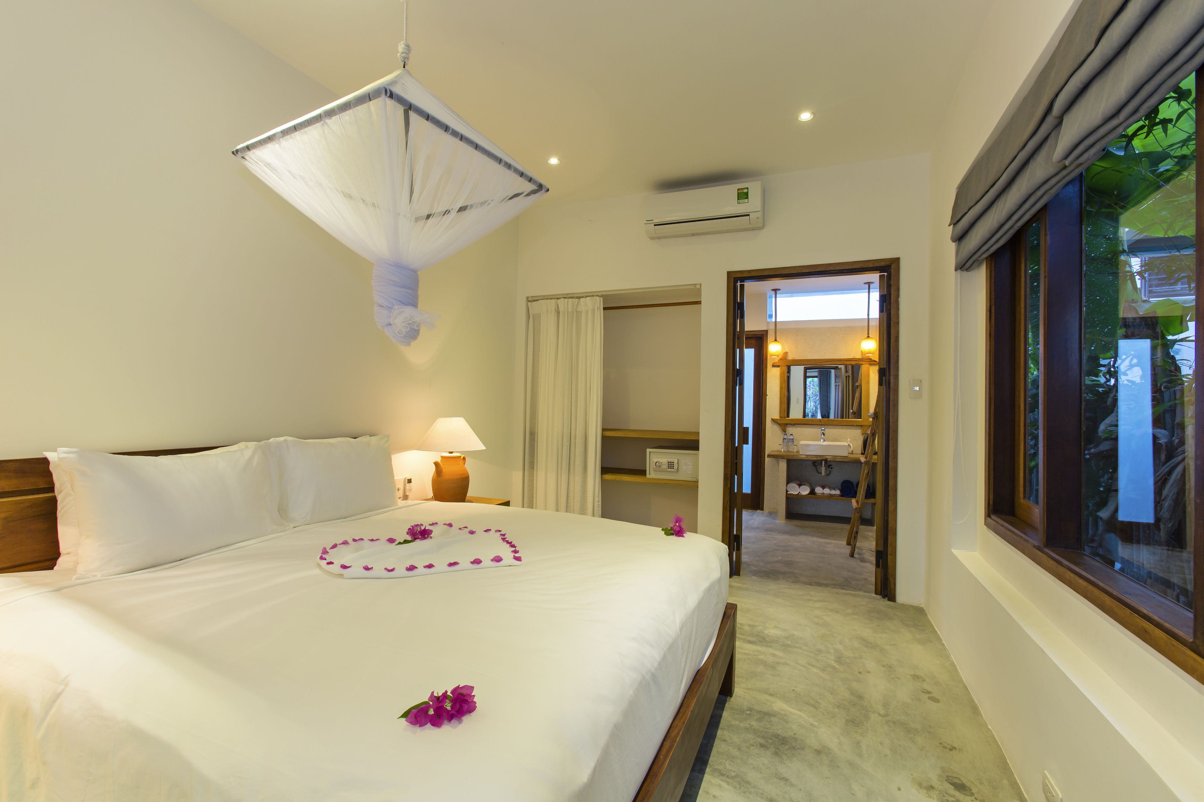 Quarto de hotel com cama, mesa de cabeceira, luminária, ar condicionado e vista para o exterior pela janela.