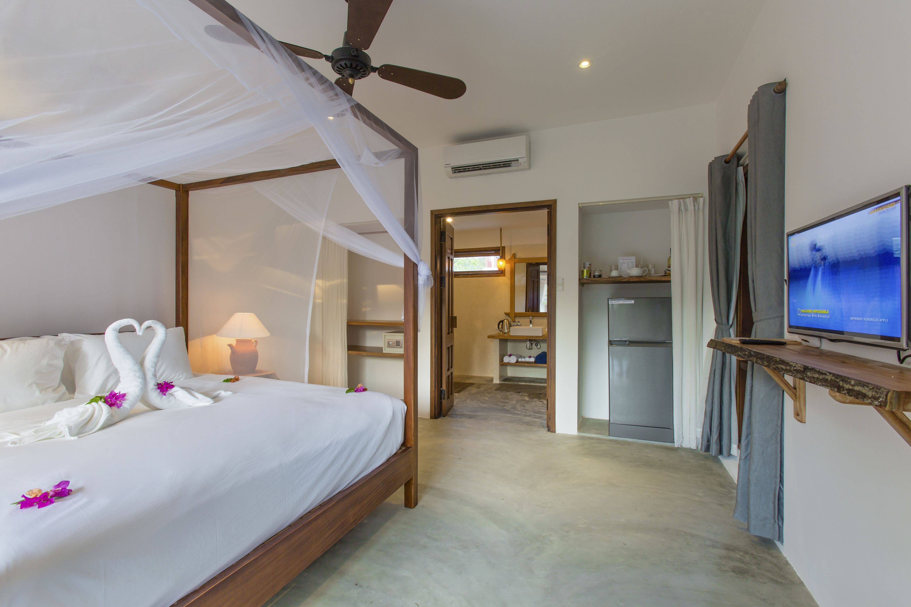 A hotel room with a four-poster bed, a ceiling fan, a television, and a bathroom visible through an open door.
