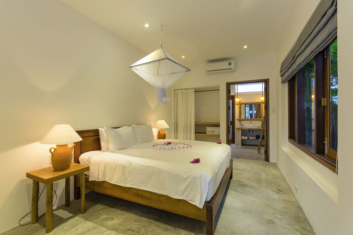 A bedroom with a bed, bedside lamps, a hanging light fixture, an air conditioning unit, a bathroom with a mirror, and a window.