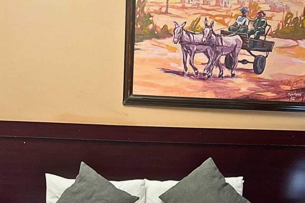 Una habitación de hotel con una cama, almohadas, una colcha y un cuadro de un carruaje tirado por caballos en la pared.
