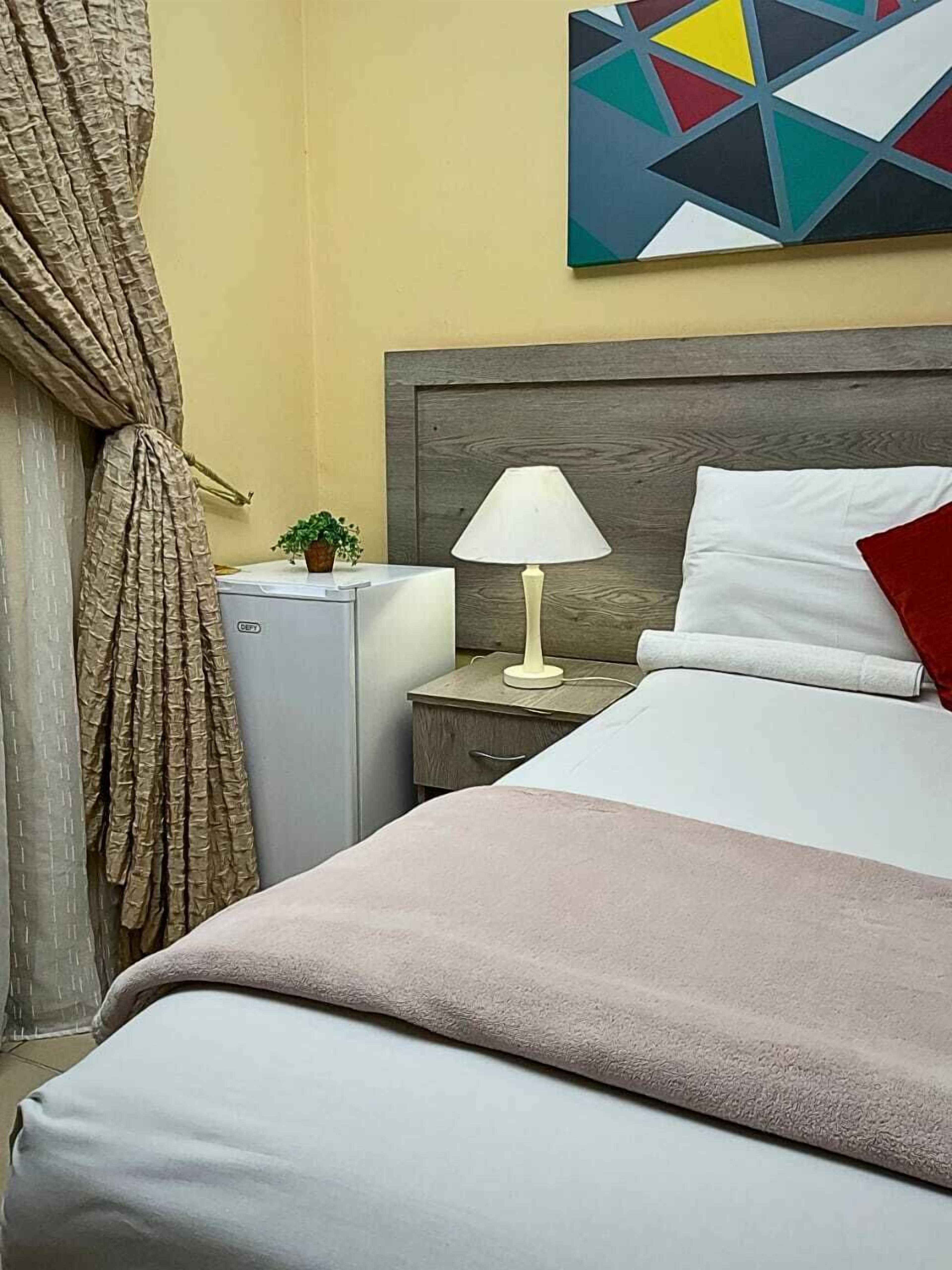 Una habitació d'hotel amb un llit, una tauleta de nit amb una làmpada, una planta petita i una obra d'art mural geomètrica.