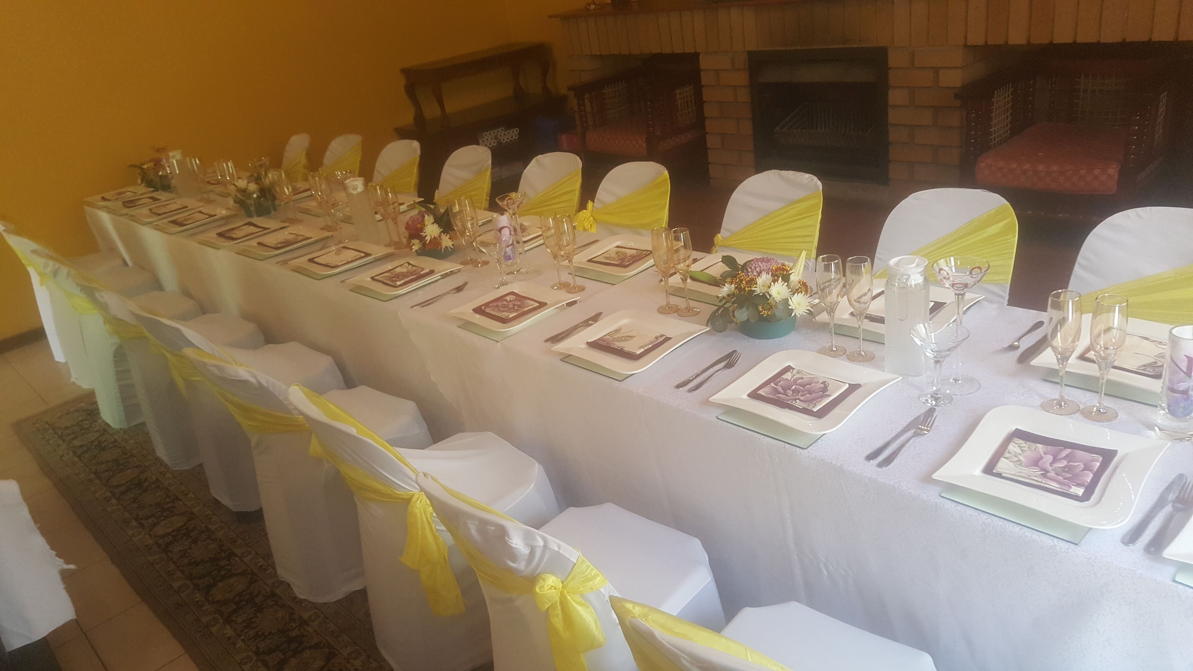 Una larga mesa de comedor dispuesta para un evento formal, con manteles blancos, fajas amarillas y centros de mesa florales.